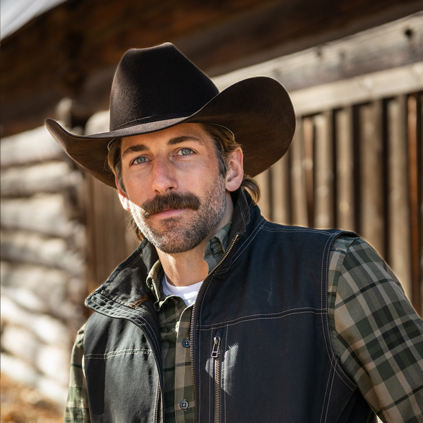 Man wearing a cowboy hat and vest in front of a wooden building