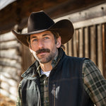 Man wearing a cowboy hat and vest in front of a wooden building