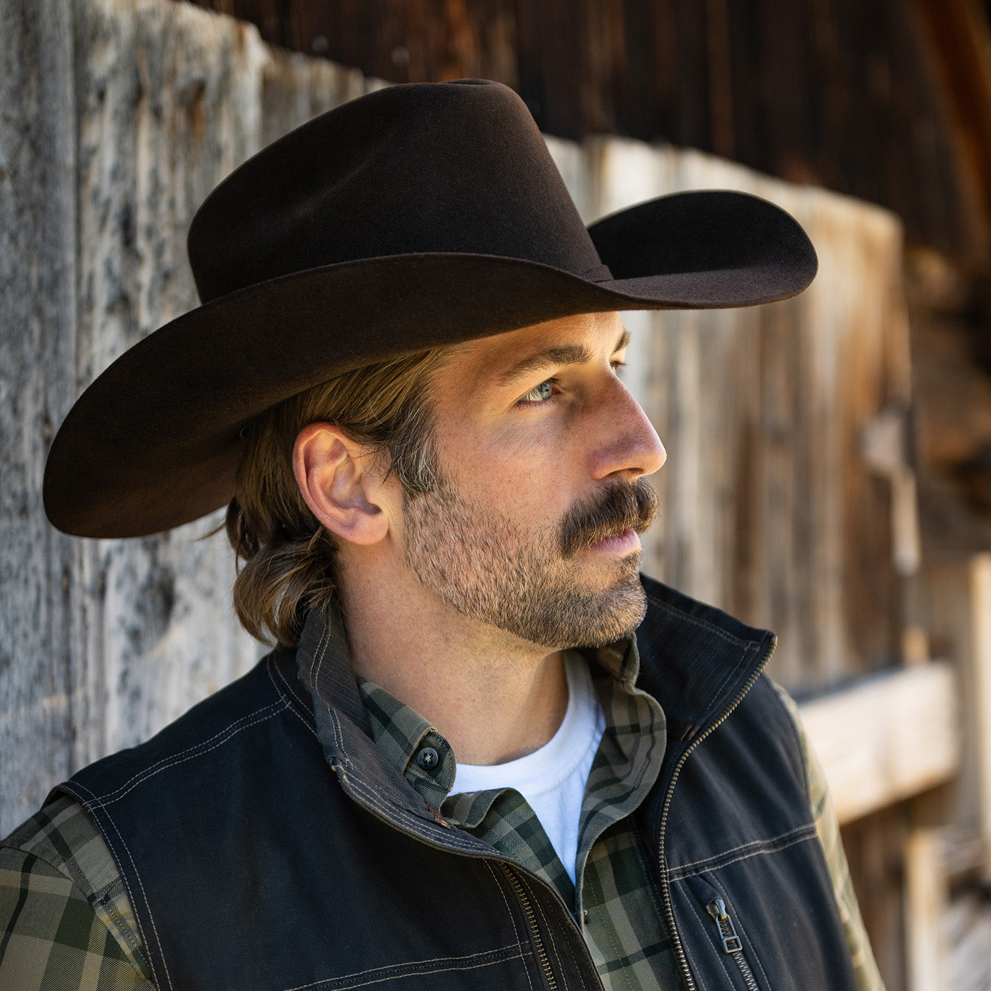 Man wearing a cowboy hat and vest standing against a wooden wall.