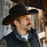 Man wearing a cowboy hat and vest standing against a wooden wall.