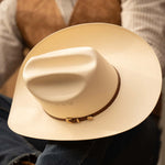 15X El Primero cognac cowboy hat resting in a person's lap with brown band and gold buckle showing the full scope of the top of the hat