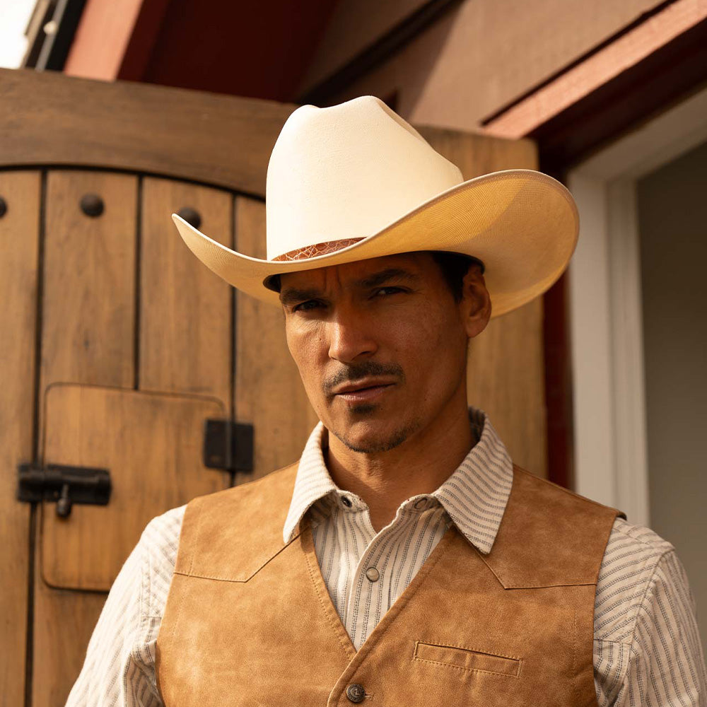 Man wearing a 15X El Primero cognac cowboy hat with brown band, vest, and white striped shirt standing near a big wooden door