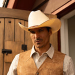 Man wearing a 15X El Primero cognac cowboy hat with brown band, vest, and white striped shirt standing near a big wooden door