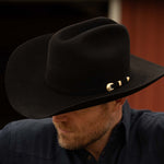 Bearded man wearing a 30X El Rey black cowboy hat and slate-colored long sleeve shirt with his head down and to the left to show the gold buckle on the brown band