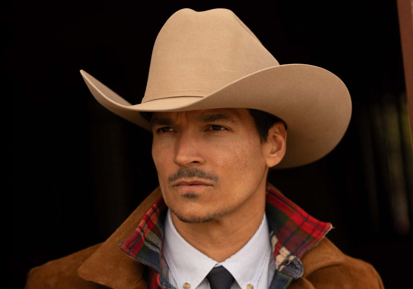 A man wearing a tan colored cowboy hat and heavy brown jacket with white undershirt and black tie