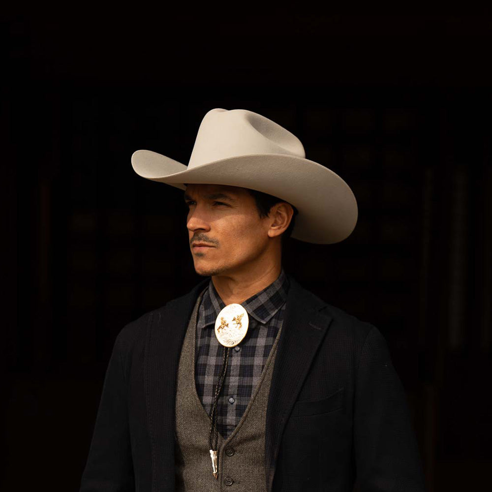 Man wearing a 100X Independencia belly cowboy hat with black jacket, grey shirt and vest, and silver bolo tie with gold horse riding emblems