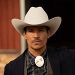 Man wearing a 100X Independencia belly cowboy hat with black jacket, grey shirt and vest, and silver bolo tie with gold horse riding emblems