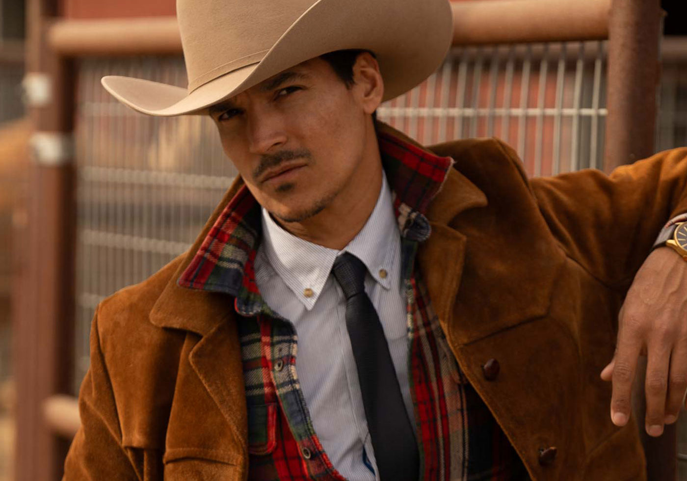 Man wearing a tan cowboy hat and brown jacket, black tie, and white shirt, and red and green plaid long sleeve shirt leaning his left arm against a wood and steel fence