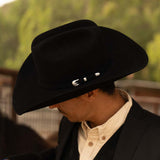Man wearing a 6X Lockhart black cowboy hat with a black jacket and white under shirt against a dark green background while turning his head to the left to show the silver buckle on the band