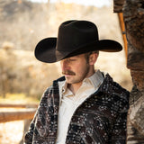 Man wearing a black cowboy hat and patterned jacket in a rustic setting