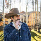 Man wearing a cowboy hat and blue patterned jacket standing in a forest with autumn foliage.