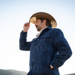 Man wearing a blue patterned coat and cowboy hat against a clear sky.
