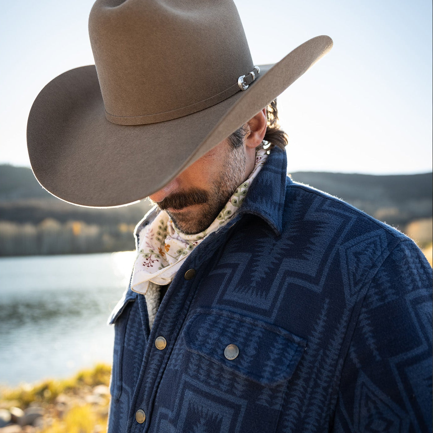 Man wearing a cowboy hat and patterned jacket by a body of water