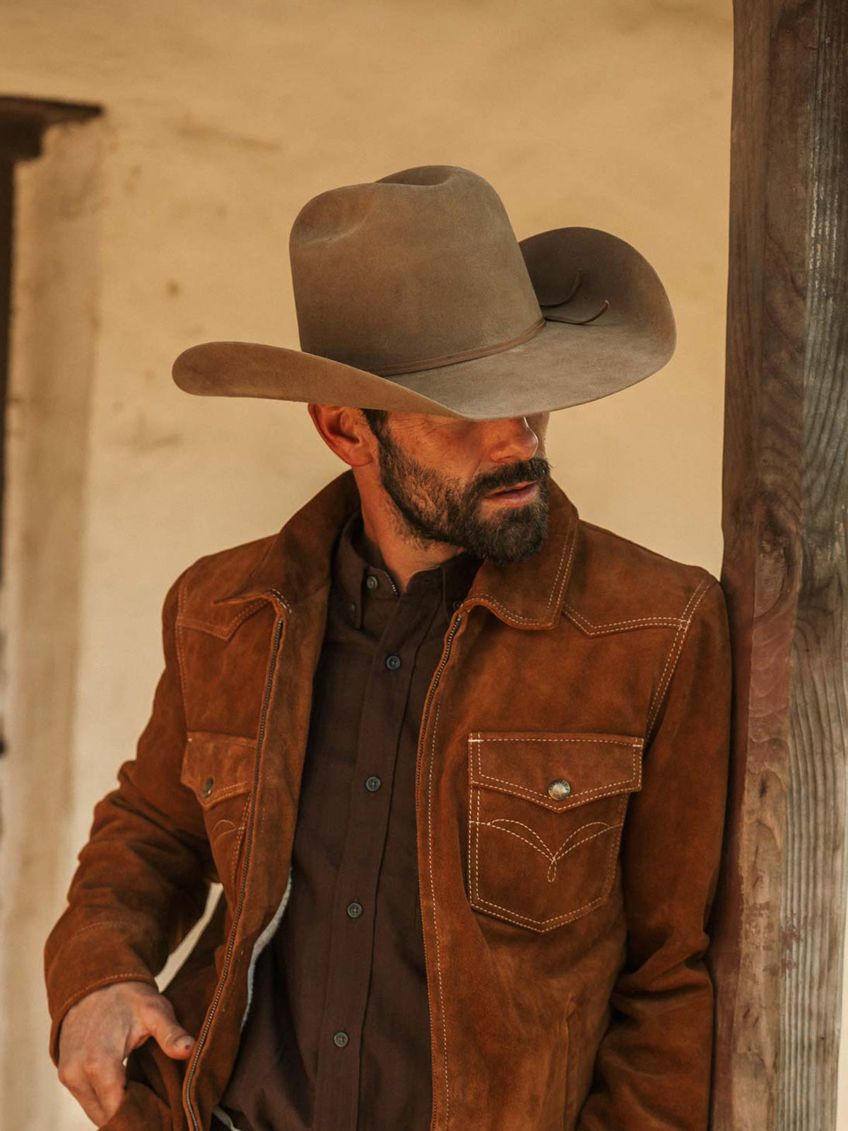 Man wearing a fawn colored cowboy hat, dusty brown jacket, and dark brown button up shirt leaning against a wooden post