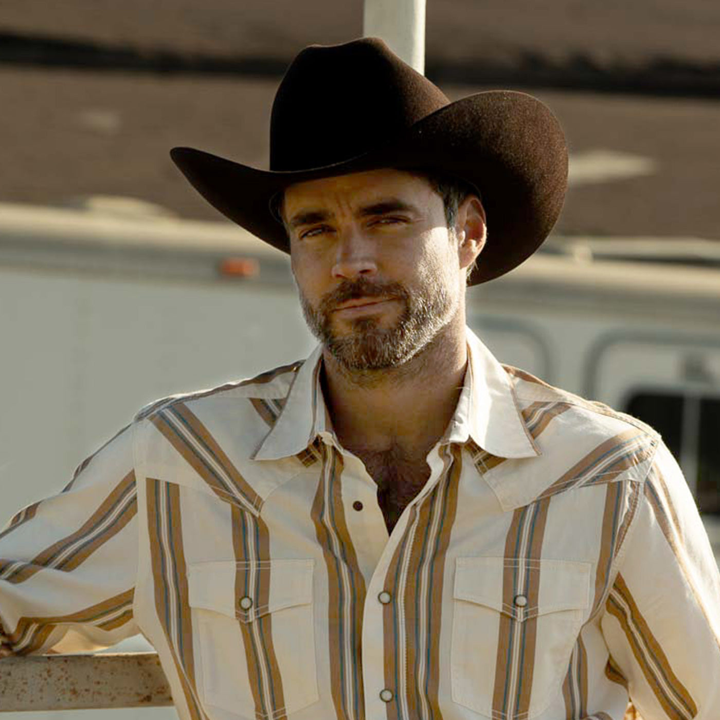 A man wearing a black cowboy hat and a faded blue, red, and white striped western shirt, photographed outdoors against a blurred rustic background