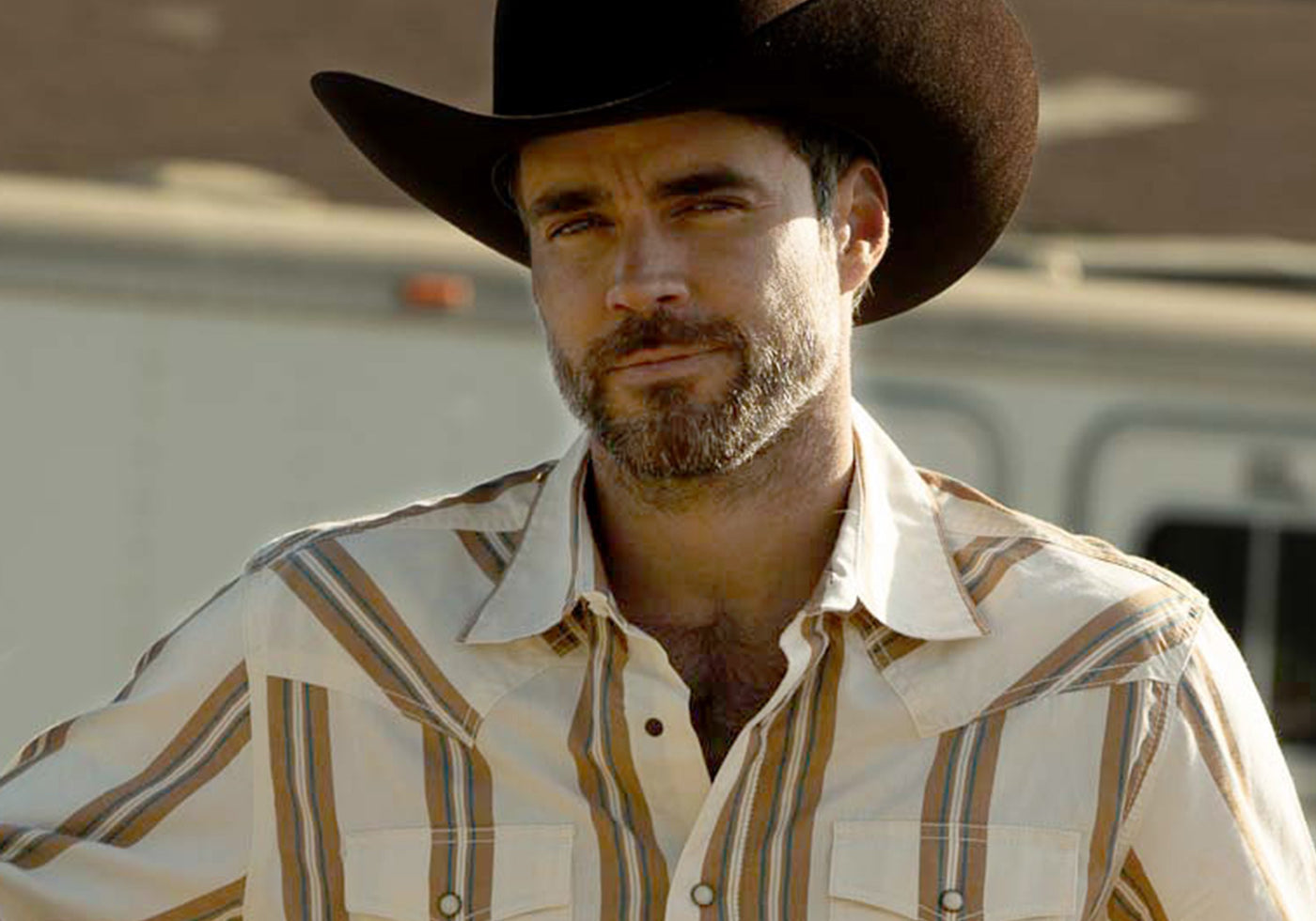 A man wearing a black cowboy hat and a faded blue, red, and white striped western shirt, photographed outdoors against a blurred rustic background