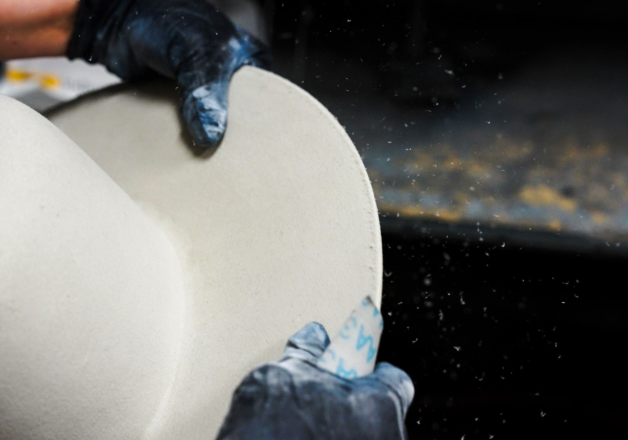 White hat brim being sanded down by a woman with black gloves as fabric bits flies off to the right