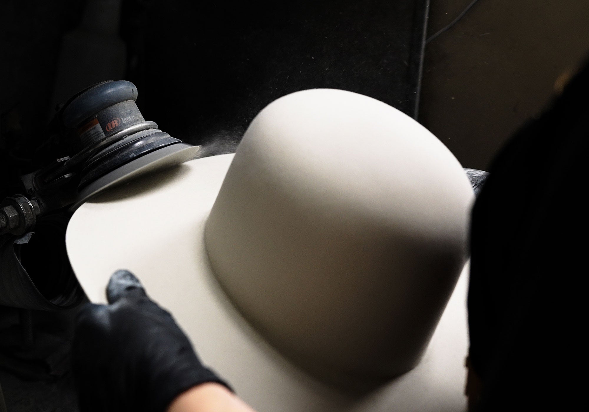White plain hat being sanded against a sanding machine held against by a person wearing black gloves