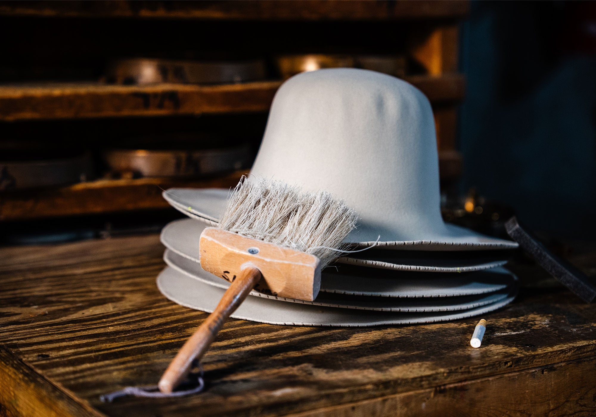 Five stacked white hat bodies on a wooden table as a white brush's bristles  rests on top of the stack with a piece of chalk and black sponge off to the right side