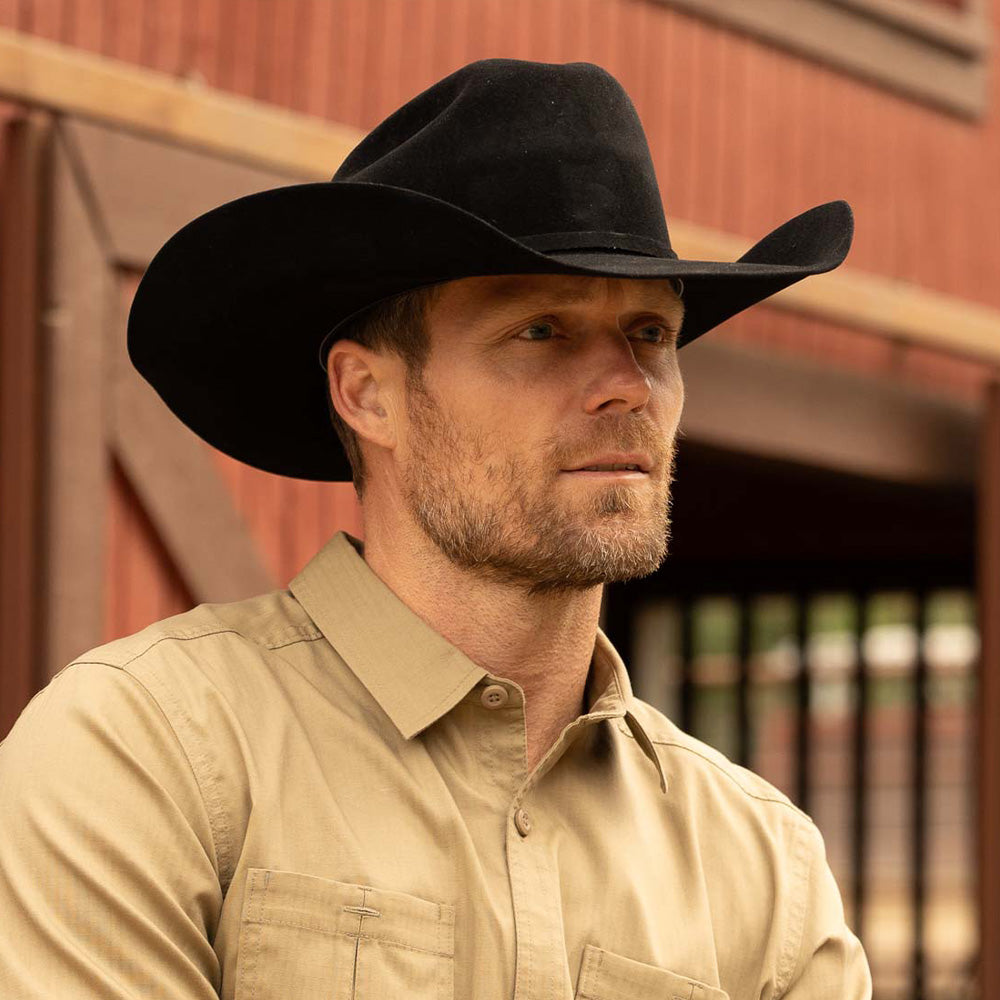 Bearded man wearing a 20X Pinnacle black cowboy hat and tan shirt standing in front of a red and brown barn