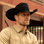 Bearded man wearing a 20X Pinnacle black cowboy hat and tan shirt standing in front of a red and brown barn