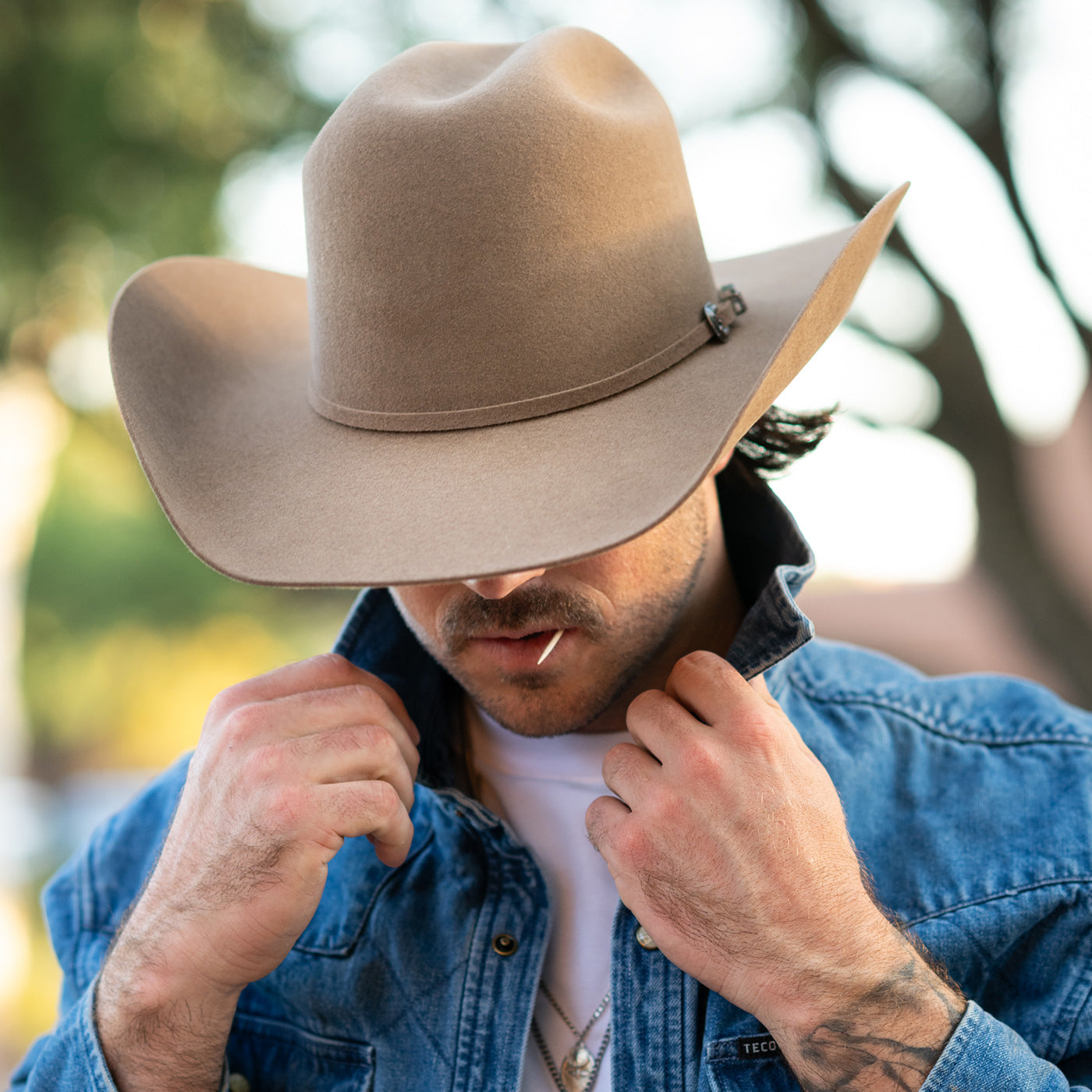 A man wearing a light brown felt cowboy hat with a small decorative pin on the band, paired with a blue denim shirt over a white t-shirt, in an outdoor setting with a blurred background