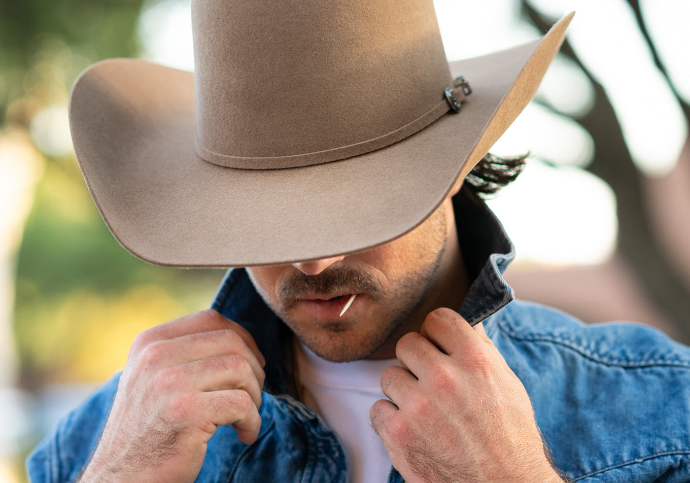 A man wearing a light brown felt cowboy hat with a small decorative pin on the band, paired with a blue denim shirt over a white t-shirt, in an outdoor setting with a blurred background